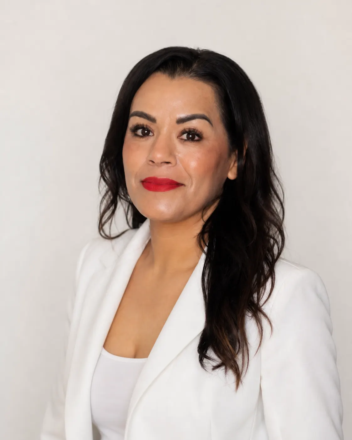 Claudia Huerta in a white blazer, looking at the camera against a light studio backdrop.